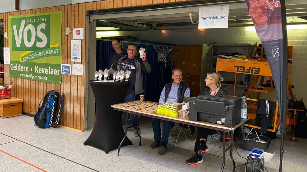 Die Turnierleitung des Badminton Cups sitzt am Meldetisch in der Halle, daneben die Pokale für die Siegerehrung