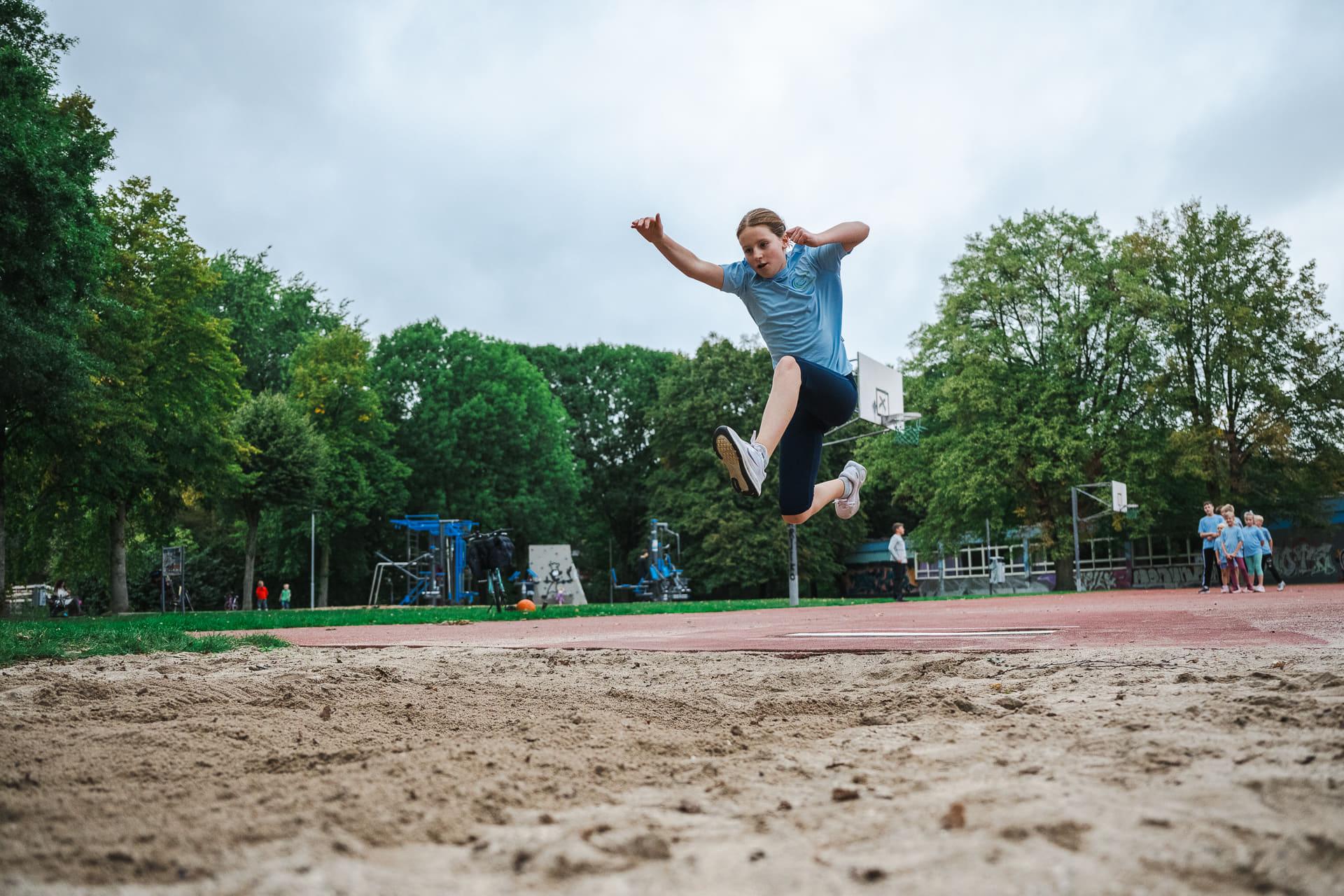 Mädchen im hellblauen Vereinsshirt fliegt beim Weitsprung mit ausgebreiteten Armen über die Sandgrube